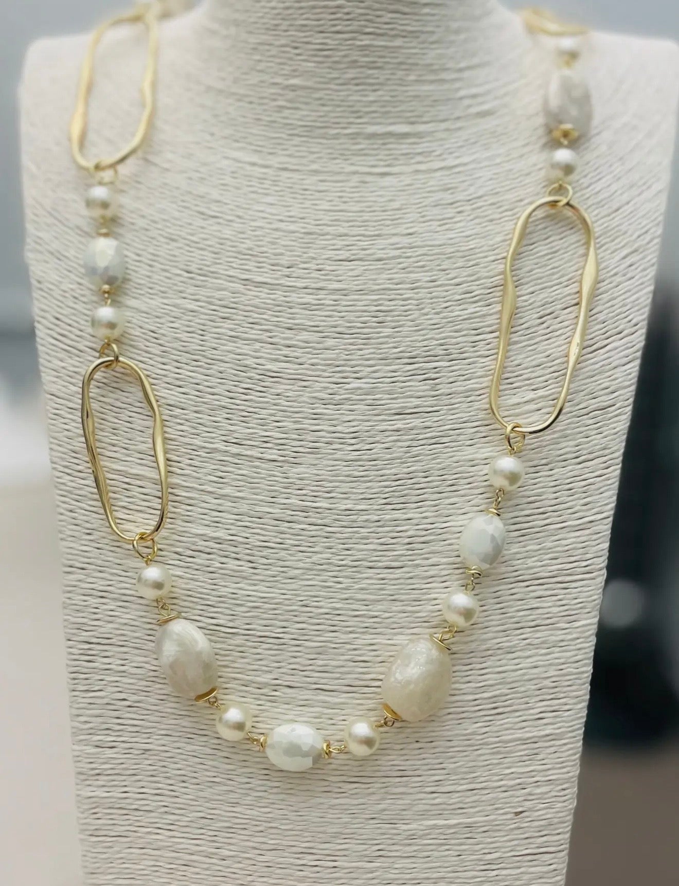 Long gold necklace with pearl beads, neutral stones, and abstract gold links displayed on a boutique jewelry stand