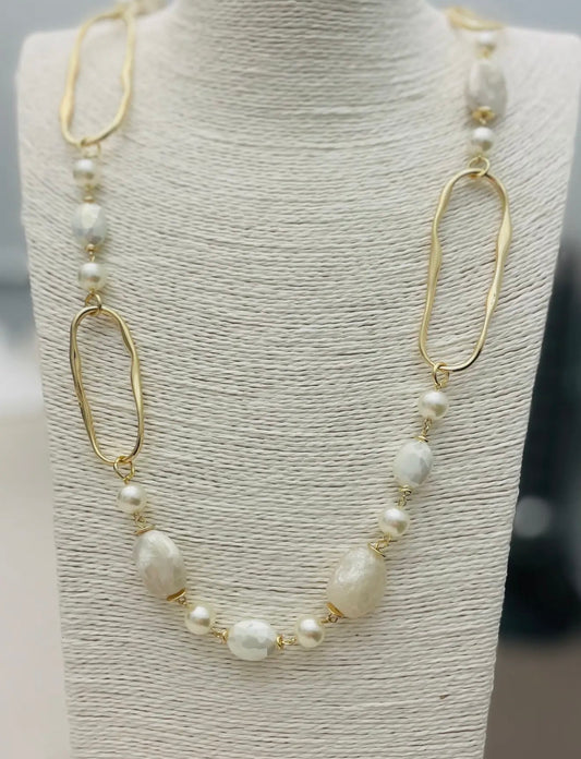 Long gold necklace with pearl beads, neutral stones, and abstract gold links displayed on a boutique jewelry stand
