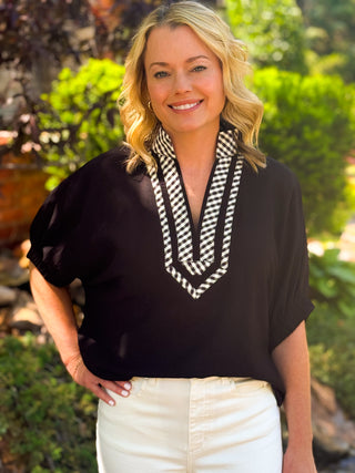Woman wearing the black Soho Lane top with contrast gingham V-neck detail and white pants outdoors.