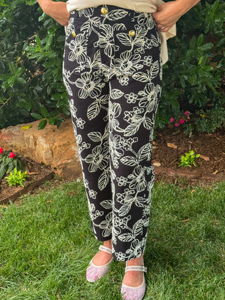 Front view of black ankle-length pants with ivory floral embroidery and straight-leg silhouette, paired with a neutral sleeveless knit top.