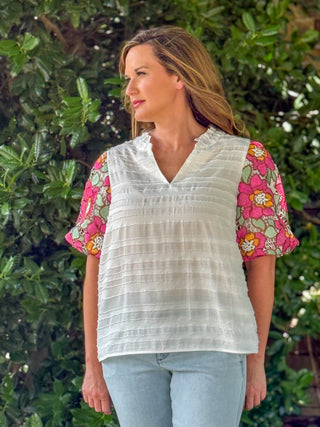 Side view of an ivory textured top with colorful floral embroidered lace sleeves in pink, sage green, and orange, paired with light wash jeans.