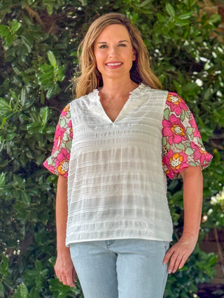 Women's ivory textured V-neck blouse featuring vibrant pink floral embroidered puff sleeves, styled with light wash denim for a casual boutique look.
