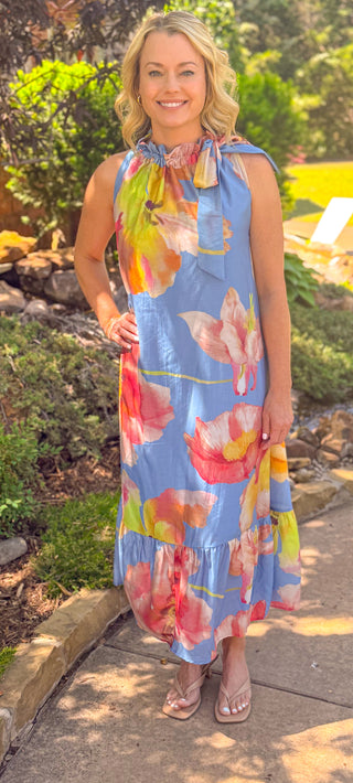 Blue sleeveless midi dress with oversized watercolor floral print, ruffle neckline, and tie shoulder detail styled outdoors.
