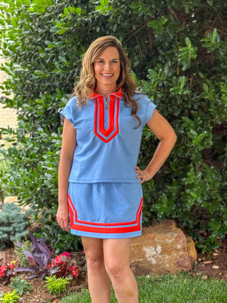 Women's blue short sleeve top and skirt set with bright red contrast trim, half-zip neckline, and coordinating mini skirt, styled outdoors for summer fashion.